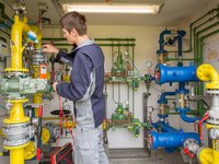 Ein junger Mann arbeitet an einer komplexen Rohrleitungsanlage. Er überprüft Geräte und Ventile an gelb, grün und blau gefärbten Rohren. Druckmessgeräte sind sichtbar, während der Mann mit einem Messgerät in der Hand beschäftigt ist.