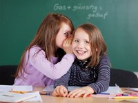Zwei lächelnde Mädchen sitzen an einem Tisch in einem Klassenzimmer. Das eine Mädchen flüstert dem anderen etwas ins Ohr. Auf dem Tisch liegen Notizblätter und Stifte. Im Hintergrund ist eine Tafel sichtbar.