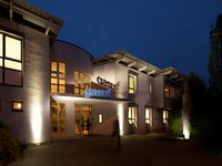 Das Bild zeigt die hell erleuchtete Fassade der gasuf bei Nacht. Über dem Eingang steht das Wort "gasuf" in blauer Schrift. Die Umgebung ist schattig, und zwei Personen sind in der Nähe des Eingangs zu sehen.