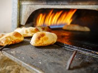 Im Bild sind frisch gebackene Teigwaren zu sehen, die aus einem Steinofen herausgenommen werden. Der Ofen ist in Betrieb, und Flammen leuchten im Hintergrund. Die Teigwaren zeigen eine goldbraune Farbe und sind auf einer schwarzen Backfläche platziert.