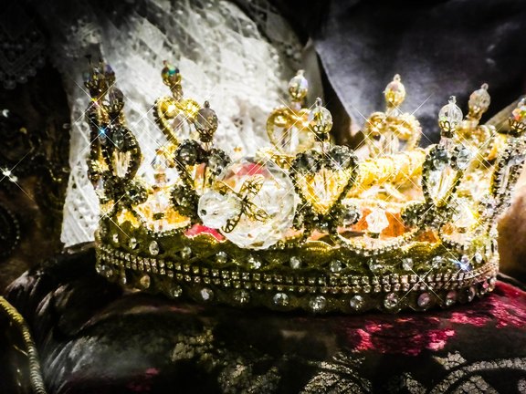 Eine opulente Krone aus goldenem Material, reich verziert mit funkelnden Edelsteinen und glitzernden Verzierungen. Die Krone hat mehrere spitz zulaufende Elemente und strahlt Eleganz und königlichen Glanz aus. Sie befindet sich auf einem stofflichen Hintergrund.