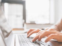 Eine Person tippt auf einer Laptop-Tastatur. Die Hände sind gut sichtbar, während der Bildschirm im Hintergrund dezent verschwommen ist. Natürliches Licht fällt durch ein Fenster und schafft eine helle, freundliche Atmosphäre.