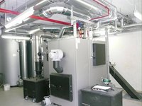 Bildbeschreibung: Der Raum zeigt eine Heizungsanlage mit mehreren silbernen Rohren, die an der Decke verlaufen. In der Mitte steht ein großer grauer Heizkessel neben einem schwarzen Behälter. Auf dem Boden liegen einige technische Geräte und Unterlagen.