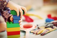 Ein Kind baut mit bunten Bauklötzen. Die Hand hält einen grünen Stein, der auf einem blauen Stein platziert wird. Die Türme bestehen aus verschiedenen Farben: grün, blau, rot und gelb. Die Umgebung ist spielerisch gestaltet.