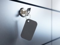 Ein Schlüssel steckt in einem Schlüsselloch, an dem ein Schlüsselanhänger mit einer grauen, rechteckigen Form befestigt ist. Die Oberfläche im Hintergrund erscheint glatt und hell, möglicherweise aus Metall oder Glas.