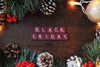 Ein Holzuntergrund mit festlichem Dekor, umgeben von Tannenzweigen und Zapfen, zeigt die Worte „BLACK FRIDAY“ in roten Scrabble-Spielsteinen. Die Buchstaben sind in zwei Reihen angeordnet und heben sich von der natürlichen Umgebung ab.