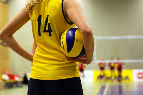 Eine Volleyballspielerin hat einen Volleyball unter den Arm geklemmt und steht am Rand eines Spielfelds
