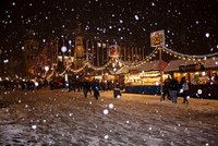 Ein Weihnachtsmarkt in der Nacht, bedeckt mit Schnee und umgeben von festlich beleuchteten Ständen. Viele Menschen schlendern über den Platz, während Schneeflocken sanft herabfallen. Lichterketten und Dekorationen schaffen eine gemütliche, festliche Atmosphäre.