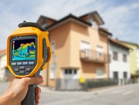 Eine Person hält ein Wärmebildgerät in der Hand, das ein farblich codiertes Wärmebild eines Hauses zeigt. Die Darstellung zeigt verschiedene Temperaturen an der Fassade, während das Hintergrundbild unscharf ist.