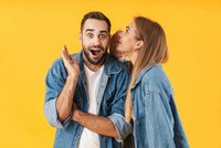 Ein überraschter Mann mit Bart trägt ein Jeanshemd und hält seine Hand an sein Gesicht. Eine Frau mit langen, blonden Haaren und ebenfalls in Jeanshemd, flüstert ihm etwas ins Ohr. Der Hintergrund ist gelb, was eine fröhliche Atmosphäre erzeugt.