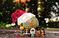Sechs farbenfrohe Holzfiguren in unterschiedlicher Kleidung stehen vor einem Globus, der eine Weihnachtsmütze trägt. Der Hintergrund ist unscharf und es scheint, als falle Schnee. Die Figuren repräsentieren verschiedene Kulturen und vermitteln eine Botschaft von Vielfalt und internationaler Gemeinschaft.