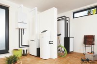 Das Bild zeigt einen modernen Raum mit Heizungsanlagen. Im Vordergrund befinden sich zwei weiße Geräte, die Heizungs- und Warmwasserfunktionen erfüllen. An der Wand verlaufen Rohrleitungen. Neben den Geräten steht ein grünes Kinderrad und im Hintergrund sind Pflanzen und Fenster zu sehen.