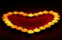 Eine Anordnung von Teelichtern in Herzform leuchtet warm auf einem roten Untergrund. Die Flammen der Kerzen strahlen ein romantisches und einladendes Licht aus.
