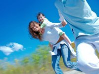 Eine fröhliche Frau mit langen, lockigen Haaren trägt ein Kind auf dem Rücken. Beide lachen und scheinen viel Spaß zu haben, während sie im Freien in Richtung Kamera laufen. Ein blauer Himmel mit einigen Wolken ist im Hintergrund sichtbar.