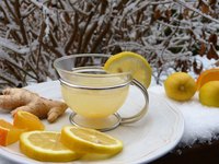 Eine hitzebeständige Glas-Teetasse mit einem gelblichen Getränk steht auf einem weißen Teller. Neben der Tasse liegen frische Ingwerstücke und mehrere Scheiben von Zitronen und Orangen. Im Hintergrund ist Schnee und verdorrtes Holz sichtbar.