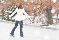 Eine Person in einem weißen Mantel und Jeans gleitet fröhlich über eine vereiste Fläche, während es schneit. Im Hintergrund sind verschwommene Pflanzen und Weihnachtslichter zu sehen.