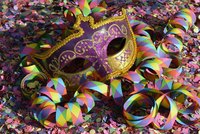 Eine bunte Maskerade-Maske in lila und golden liegt auf einem bunten Konfetti-Haufen. Rund um die Maske sind spiralförmige, farbige Papierschlangen verteilt, die für eine festliche Stimmung sorgen.