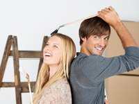 Ein lächelndes Paar steht Rücken an Rücken, beide mit Malerpinseln in der Hand. Der Mann hält seinen Pinsel über dem Kopf der Frau. Sie wirken entspannt und glücklich in einer hellen Umgebung, die eine kreative Atmosphäre vermittelt.