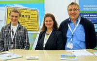 Team auf der Mainfranken-Messe steht am Informationsstand. Im Hintergrund ist ein Ortsschild von Würzburg sichtbar. Die Person links trägt ein kariertes Hemd, die in der Mitte einen blauen Blazer. Die Person rechts hat eine Brille auf und trägt einen dunklen Pullover. Auf dem Tisch liegen Informationsblätter.