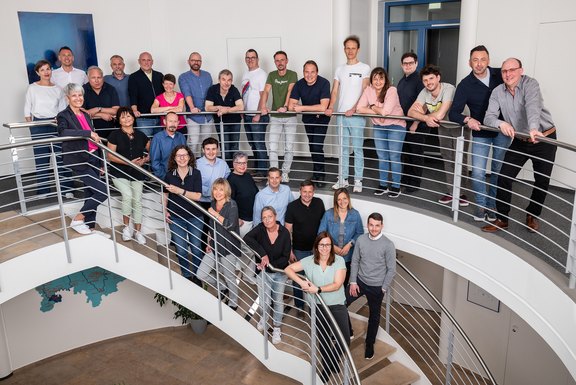 Teamfoto der gasuf-Mitarbeiter stehend auf einer geschwungenen Treppe im Bürogebäude. Sie sind in zwei Reihen angeordnet, lächeln und schauen in die Kamera. Im Hintergrund sind Pflanzen und große Fenster sichtbar.
