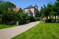 Ein gepflasterter Weg führt durch eine grüne Wiese nach Bastheim Vorbei an einem historischen Gebäude mit einem Glockenturm. Umgeben von Bäumen und einer Holzlattenzaun vermittelt die Szene eine ruhige, ländliche Atmosphäre an einem sonnigen Tag.