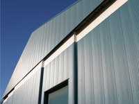 Eine moderne Gebäudeaußenwand aus vertikalen, metallischen Paneelen, die in verschiedenen Blautönen schimmert. Die Wand hat große, rechteckige Fenster und ist vor einem klaren, blauen Himmel abgebildet. Das Licht fällt gleichmäßig auf die Oberfläche und betont die Struktur.