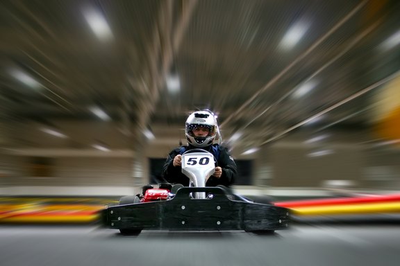 Ein Kind sitzt in einem Go-Kart mit der Nummer 50. Es trägt einen schwarzen Overall und einen Helm. Das Bild vermittelt ein Gefühl von Geschwindigkeit, da der Hintergrund verschwommen ist. Die Szenerie scheint sich in einer Innenrennbahn zu befinden.