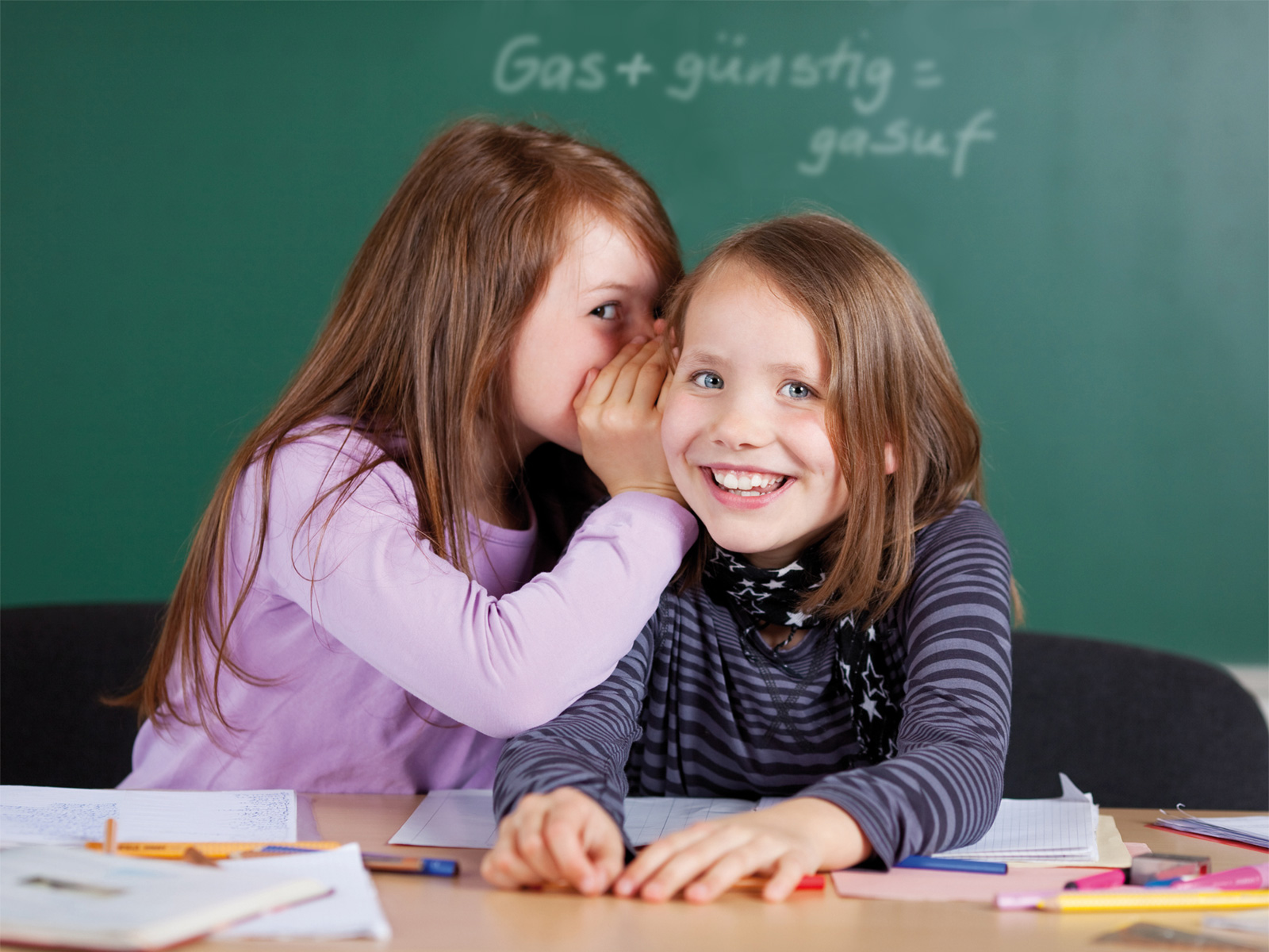 Zwei lächelnde Mädchen sitzen an einem Tisch in einem Klassenzimmer. Das eine Mädchen flüstert dem anderen etwas ins Ohr. Auf dem Tisch liegen Notizblätter und Stifte. Im Hintergrund ist eine Tafel sichtbar.